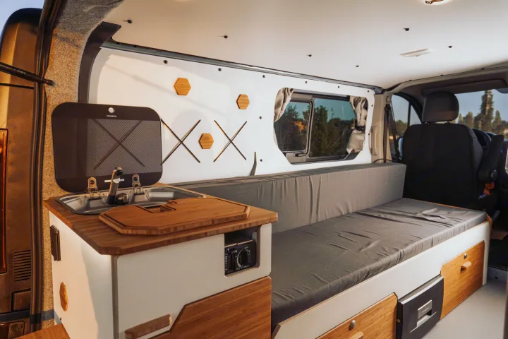 Modern rv rental interior: compact kitchen with Dometic sink, cooktop, wooden counter, and a convertible bench-bed.. Renault Trafic 2025