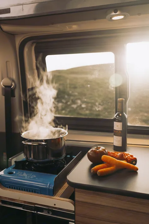 Inside an RV rental, a pot boils on a portable stove, with fresh vegetables and wine ready for a meal against a scenic nature backdrop.. Volkswagen Transporter T6.1 2022