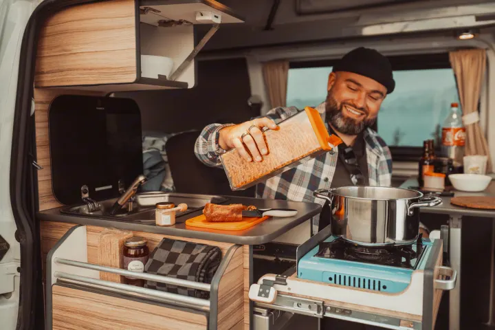 Happy man cooks pasta in his RV rental campervan, pouring into a pot on a stove in the compact kitchen.. Volkswagen Transporter T6.1 2022