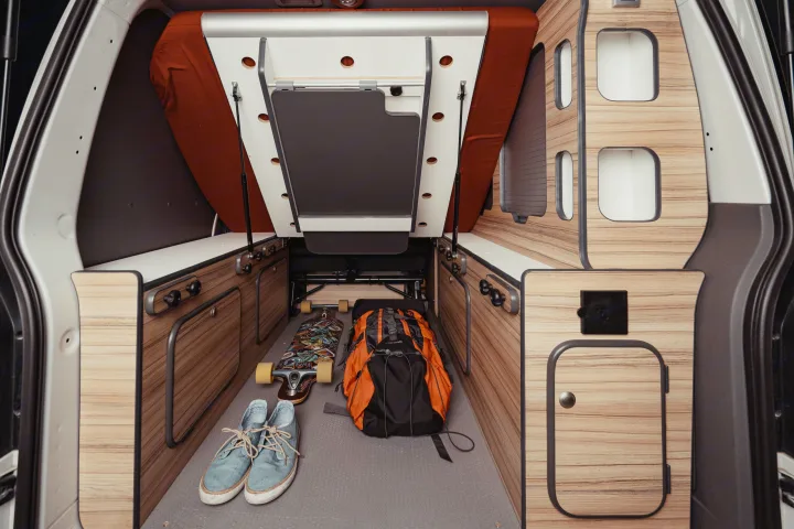 Compact RV rental interior with folded bed, wooden storage, longboard, and backpack for adventure travel.. Volkswagen Transporter T6.1 2022