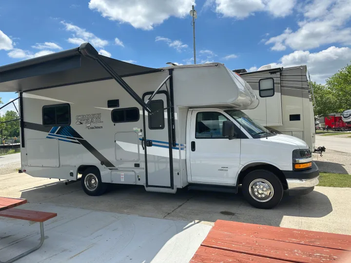 White & gray Class C rv rental (Coachmen Cross Trail) with awning extended, parked on concrete at a sunny campsite. Picnic tables visible.. Coachmen Other 2022
