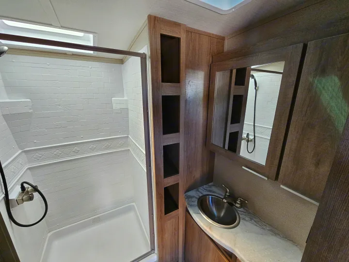 Modern RV rental bathroom, showcasing a white textured shower enclosure with glass door and a wood vanity with sink and mirror.. Grand Design Imagine 2021