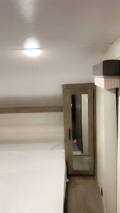 Clean RV rental bedroom with white sheets, a ceiling light, wood paneling, and a mirrored wardrobe entry.. Forest River Wildwood X-Lite 2021