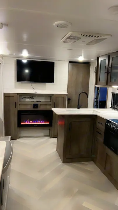 Modern RV rental interior with a compact kitchen, electric fireplace, TV, and stylish herringbone flooring for cozy travel.. Forest River Wildwood X-Lite 2021