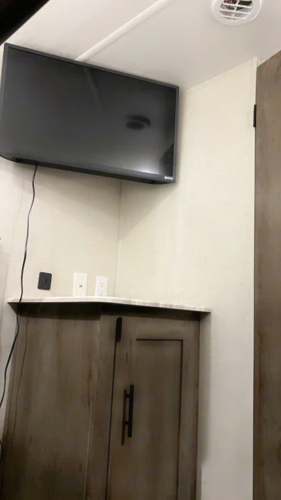Modern RV rental interior features a wall-mounted TV over a countertop and cabinet. Outlets and a ceiling vent are also visible.. Forest River Wildwood X-Lite 2021