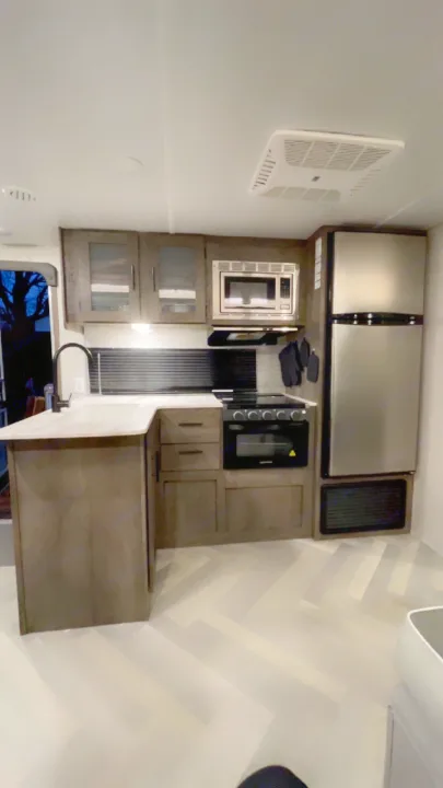 Modern RV rental kitchen with light wood cabinetry, stainless steel appliances, light countertops, and herringbone flooring.. Forest River Wildwood X-Lite 2021