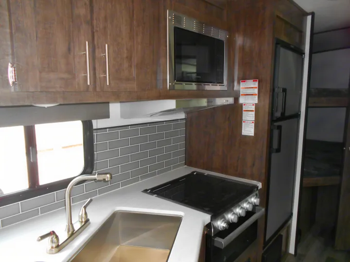 Modern RV rental kitchen interior with wood cabinets, stainless microwave, stove, sink, and large fridge. Bunk beds beyond.. Xplorer Xplorer 240BHX 2023