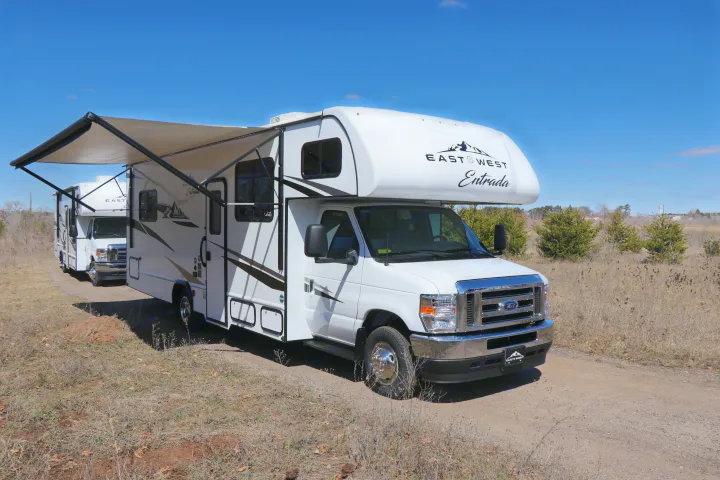 A white East West Entrada RV rental with an extended awning sits on a dirt path, another RV behind, under a clear sky.. East to West entrada 2025