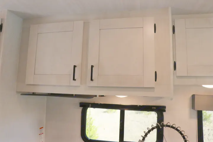 Overhead light-colored cabinets with black handles in an RV rental kitchen. Below are under-cabinet lighting, a window, and a coiled faucet.. East to West entrada 2025