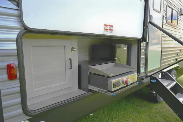 An RV rental's exterior storage bay reveals a pull-out griddle, mini-fridge, and cabinet with a bottle opener for outdoor cooking.. Forest River Aurora 28BHS 2024