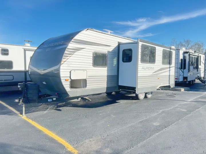 A grey and white Forest River Aurora **rv rental** travel trailer is parked on an asphalt lot under a clear blue sky.. Forest River Aurora 28BHS 2024