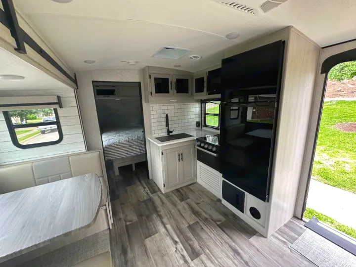 Modern RV rental interior: kitchen with dark appliances, light cabinets, dining booth, and bedroom glimpse. Wood-look flooring.. Crossroads RV Sunset Trail Superlite 2024