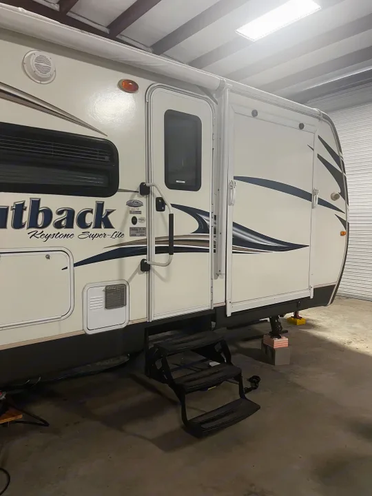 Side view of a white Keystone Outback Super-Lite travel trailer, suitable for an RV rental. Features steps & handrail.. Keystone RV Outback Toy Hauler 2013