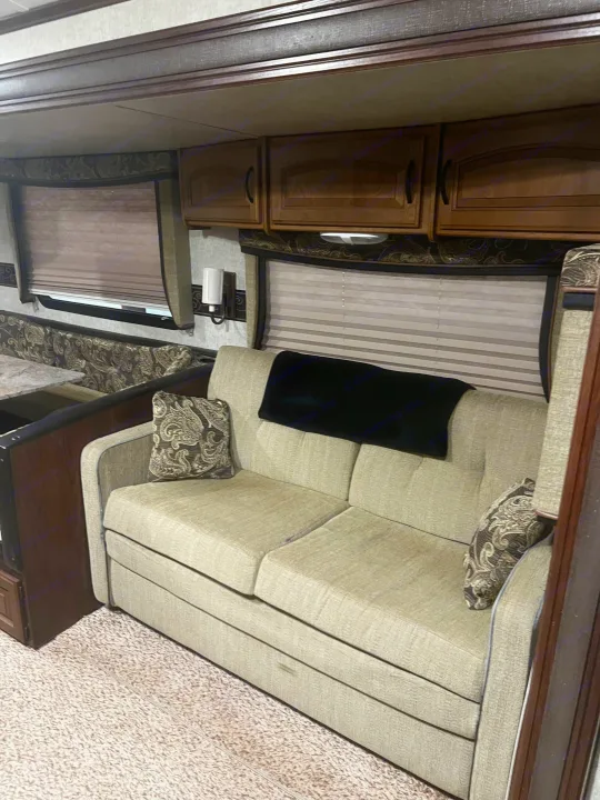 RV rental interior: A light-colored, pull-out sofa with patterned pillows, overhead cabinets, and windows with blinds.. Keystone RV Outback Toy Hauler 2013