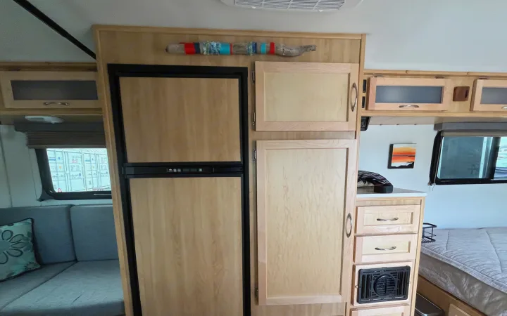 Inside an RV rental, light wood fridge, cabinets, and a dinette are visible. A window reveals shipping containers outside.. Escape Trailer Escape Trailer Trailer 2022