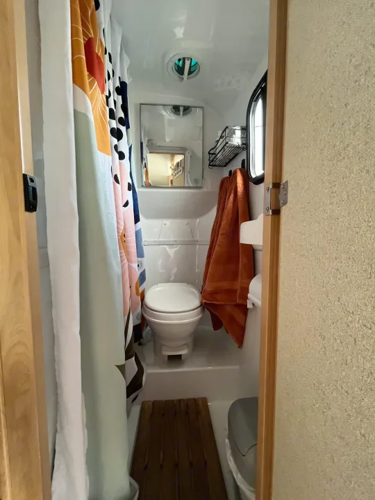 Inside a compact RV rental bathroom: toilet, shower area with patterned curtain, mirror, and fan. Ready for travel.. Escape Trailer Escape Trailer Trailer 2022