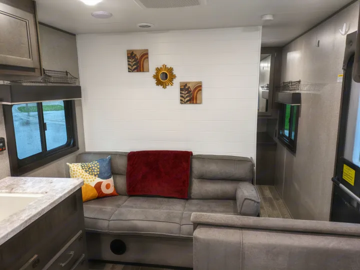 Cozy RV rental living space with a grey sofa, red throw, colorful pillow, white shiplap wall, and outdoor view through windows.. Jayco Jay Flight SLX 2021