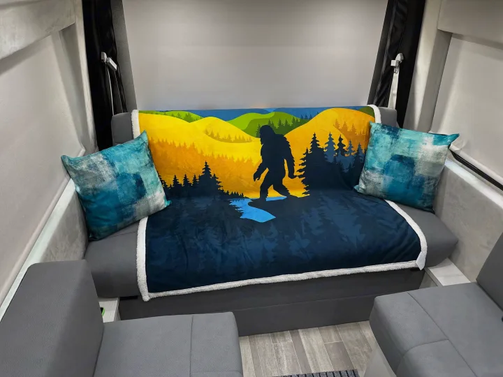 Inside an RV rental, a bed/couch with a dark blue Bigfoot blanket, colorful landscape, and blue pillows.. Pleasure-Way Pleasure-Way Class B 2024