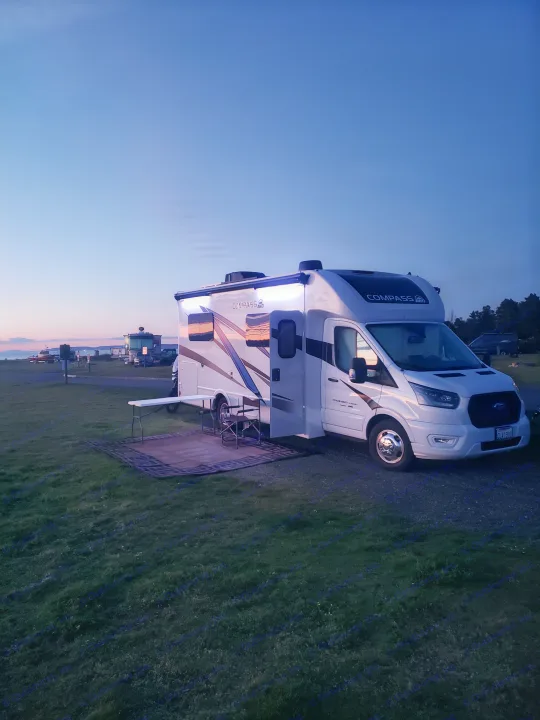 RV rental at a scenic campsite. An illuminated motorhome is parked on grass at dusk with a table and mat.. Thor Compass 2024
