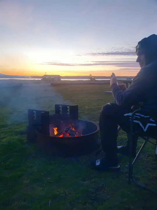RV rental adventure: Person relaxes by a glowing campfire, watching a beautiful sunset over the waterfront.. Thor Compass 2024