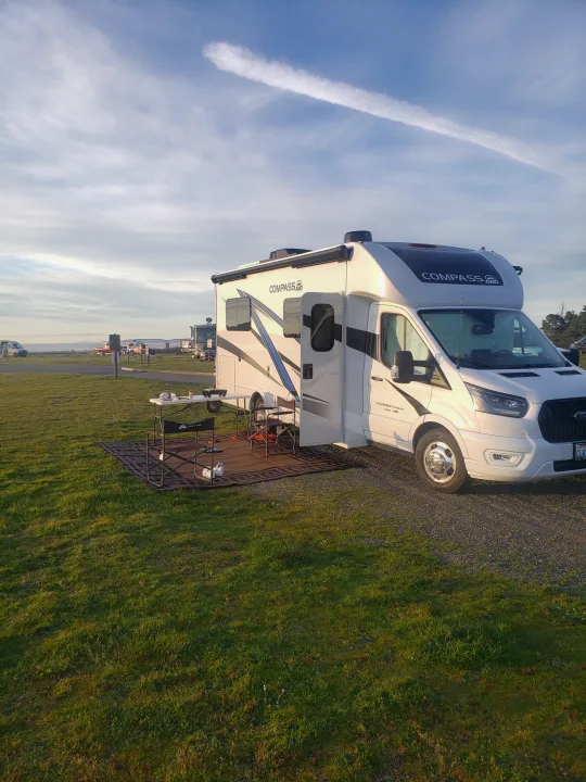 A white Compass RV rental is parked at a grassy waterfront campsite with an outdoor table & chairs.. Thor Compass 2024