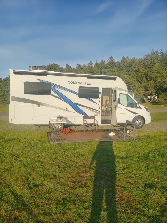 White RV rental at a sunny campsite with outdoor seating, a dog visible inside. Trees line the background.. Thor Compass 2024