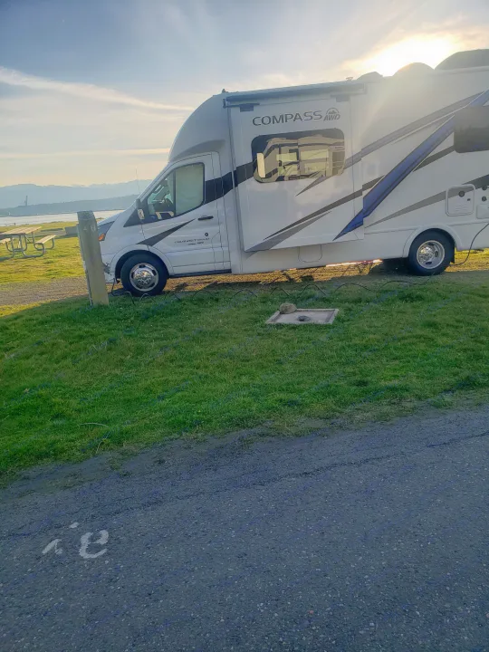 A white Compass AWD motorhome rental is parked at a scenic campsite with mountains and water, bathed in sunlight.. Thor Compass 2024