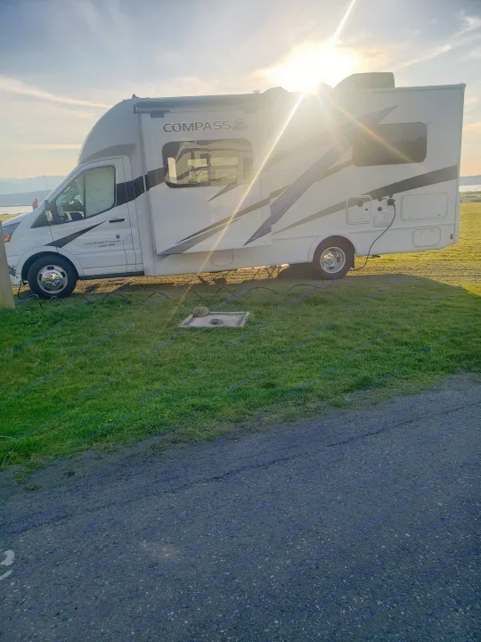 A white RV rental is parked at a scenic campsite by the water and mountains, bathed in warm sunset light.. Thor Compass 2024