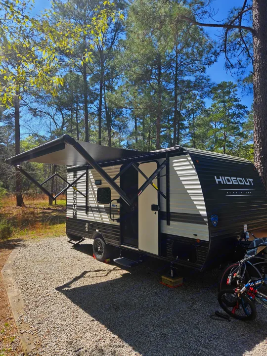 An RV rental parked on a gravel campsite with its awning extended, surrounded by tall pine trees under a clear blue sky.. Keystone RV Keystone Hideout 175BH 2024