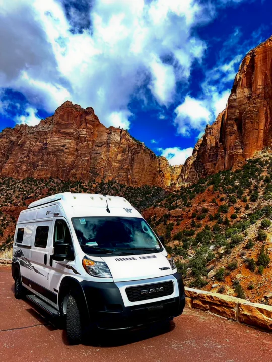 Explore stunning landscapes with an RV rental! White camper van parked by massive red rock cliffs under a dramatic blue sky.. Winnebago Solis 2024