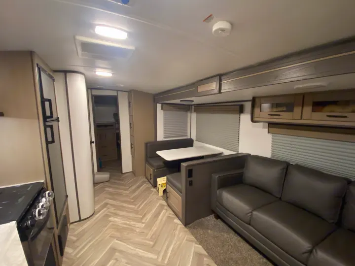 Modern RV rental living space: comfy dinette, dark sofa, kitchen with stove, and stylish light wood herringbone flooring.. Cruiser RV Shadow Cruiser 2021