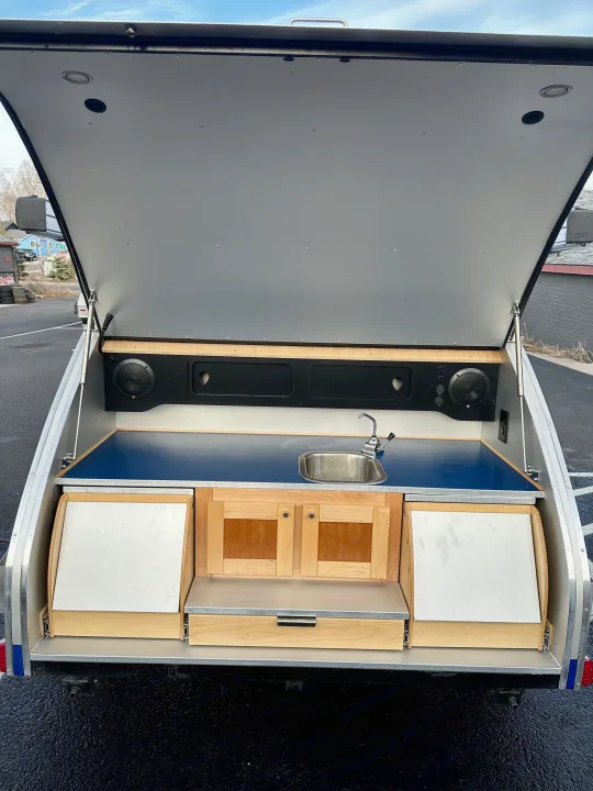 This RV rental teardrop trailer shows its open rear hatch revealing a full outdoor kitchen with sink, cabinets, and speakers.. Pindrop Travel Trailer Fina Roma 2022
