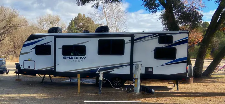 A white Shadow Cruiser RV is parked in a treed campground site with visible power hookup. Perfect for an RV rental getaway.. Cruiser RV Shadow Cruiser 2021