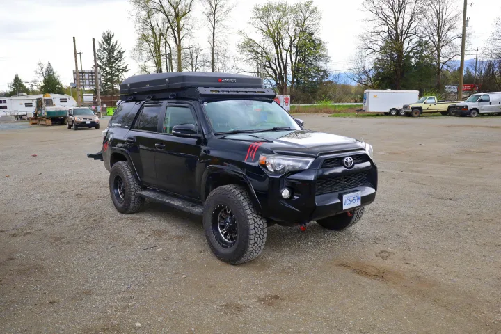 Black Toyota 4Runner with rooftop tent on gravel. Nearby: excavator, trailers & vehicles, likely an RV rental or storage lot.. Toyota Other 2015