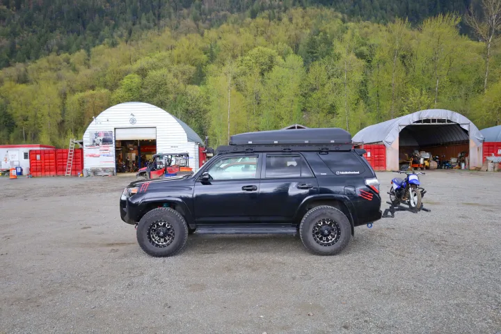 Black overlanding Toyota 4Runner with roof box & dirt bike on gravel. Great alternative to RV rental for rugged adventures.. Toyota Other 2015