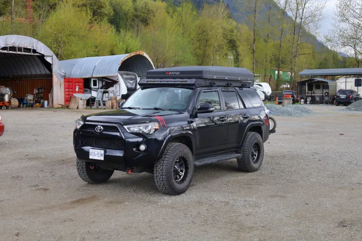 A black Toyota 4Runner with a rooftop tent on a gravel lot. Several RVs and covered storage structures suggest an rv rental or storage facility.. Toyota Other 2015