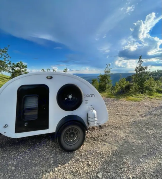 Adventure awaits! A white teardrop camper, ideal for an RV rental, is set on a gravel path overlooking a vast mountain view.. Bean Bean Stock 2021