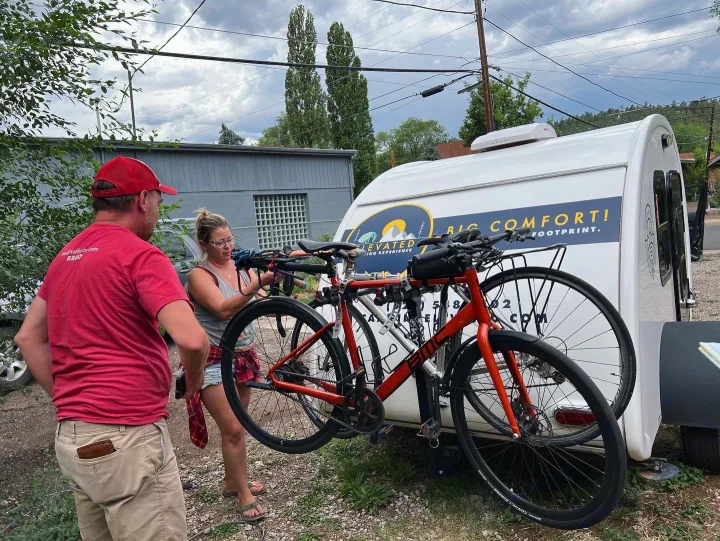 Setting up for adventure: Two people with bikes attached to a white teardrop rv rental, featuring "BIG COMFORT!" branding.. Bean Bean Stock 2021