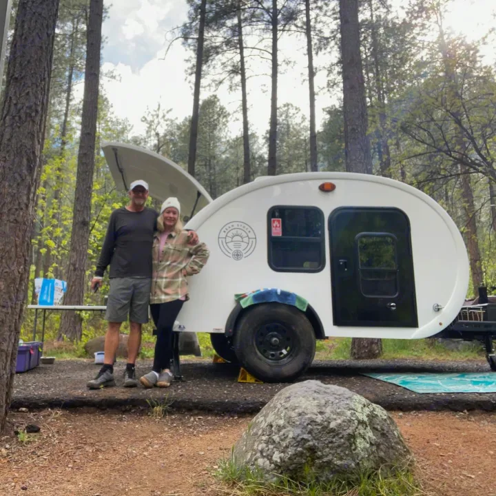 A smiling couple stands by their white teardrop RV rental in a serene pine forest campsite.. Bean Bean Stock 2021