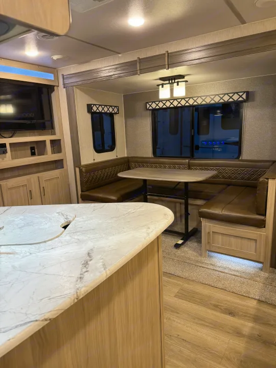 An rv rental interior featuring a white marbled kitchen island, brown U-shaped dinette, and entertainment center with natural wood.. Coachmen Freedom Express 2020