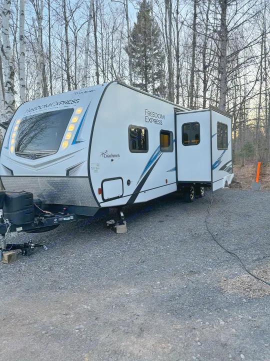White "Freedom Express" RV rental set up on a gravel campsite among bare trees. Ideal for a nature camping getaway.. Coachmen Freedom Express 2020