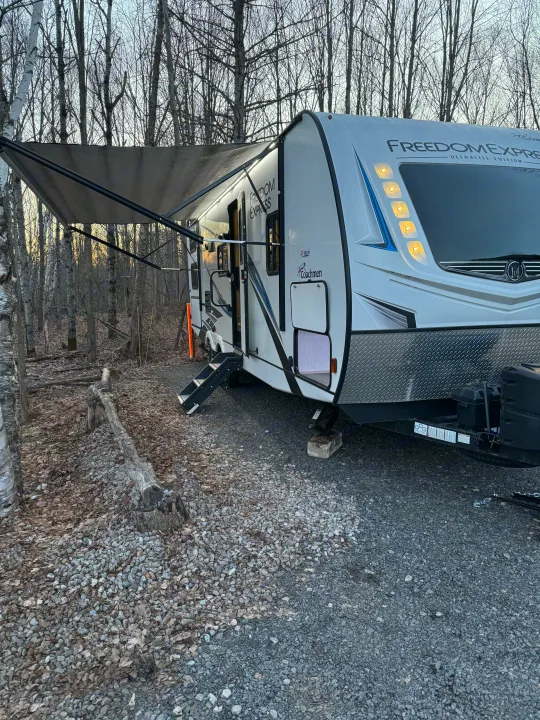 RV rental at a scenic campsite. White Freedom Express travel trailer with illuminated awning, surrounded by bare trees & gravel.. Coachmen Freedom Express 2020