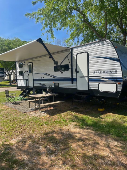 A white RV rental travel trailer at a sunny campsite with extended awning, picnic table, and grill.. Keystone RV Springdale 2021