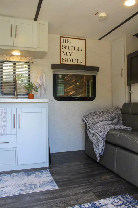 Cozy RV rental interior with a light kitchen, textured wall with "BE STILL MY SOUL" sign, and a gray sofa.. Keystone RV Bullet Crossfire 2021