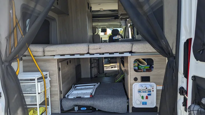 Spacious RV rental interior from the rear, showing a raised bed, storage, and utility panel for compact camper van living.. Winnebago Solis 59PX 2023