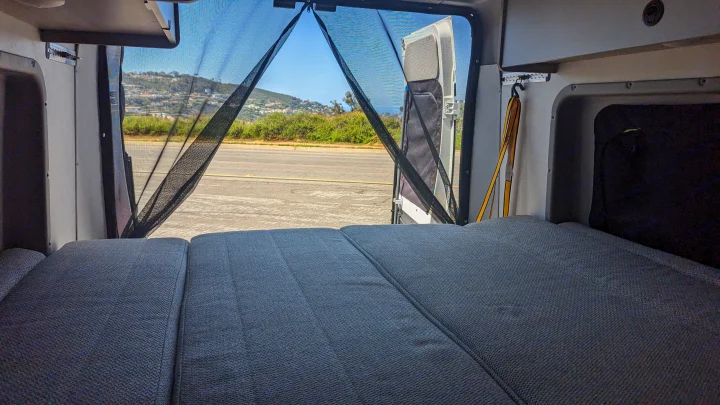 Inside a spacious RV rental, a comfortable bed faces open rear doors with a mesh screen & scenic outdoor view.. Winnebago Solis 59PX 2023