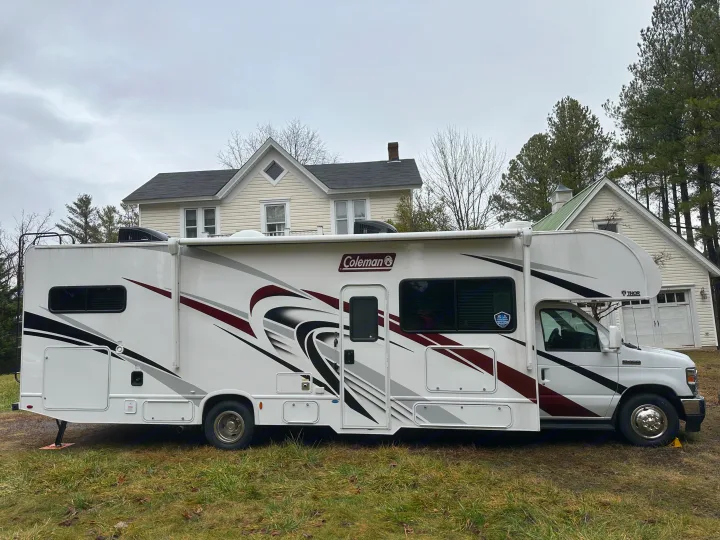 A white Coleman RV rental with red/black graphics is parked on a grassy lawn. Houses & trees form the background.. Thor Coleman 2023