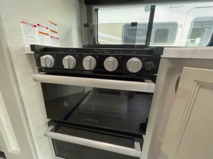 Inside an RV rental: a black Furrion stove, oven, and lower storage. Warning labels line the textured wall.. Thor Coleman 2023