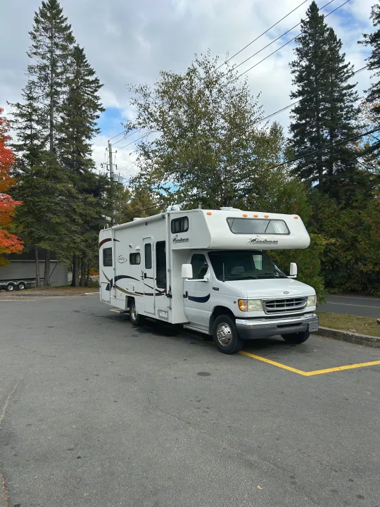 White Coachmen Class C RV rental parked in an asphalt lot with trees and an overcast sky, ready for adventure.. Coachmen Catalina 2003