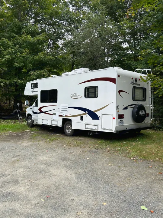 A white Coachmen Catalina Sport motorhome parked on a gravel lot amidst green trees. Perfect RV rental for camping adventures.. Coachmen Catalina 2003
